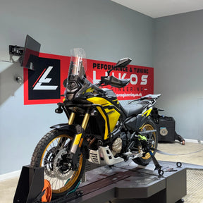 A yellow and black BMW adventure motorcycle is photographed on a dynamo stand inside a workshop. The background features a large red and black sign for 'Lukos Performance & Tuning'.